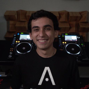 Joven sonriendo, vestido con una camiseta negra que tiene un logo blanco en el centro. Detrás de él, se observan equipos de DJ, incluyendo consolas de mezclas y otros dispositivos electrónicos.